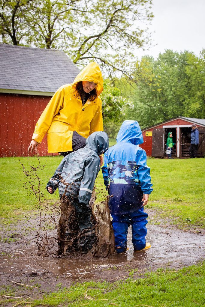 A teacher in a bright yellow rain coat stomping in rain and mud puddles with two young children in blue rain gear at The Children's Farm.