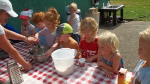 Children gathered around a table helping to measure and mix ingredients to make zucchini muffins as a snack.